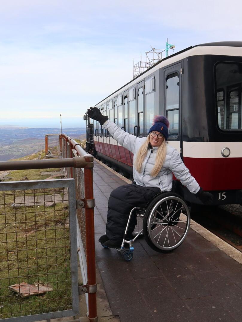 A lady in a wheelchair with arms outstretched by a train at the top of a mountain.