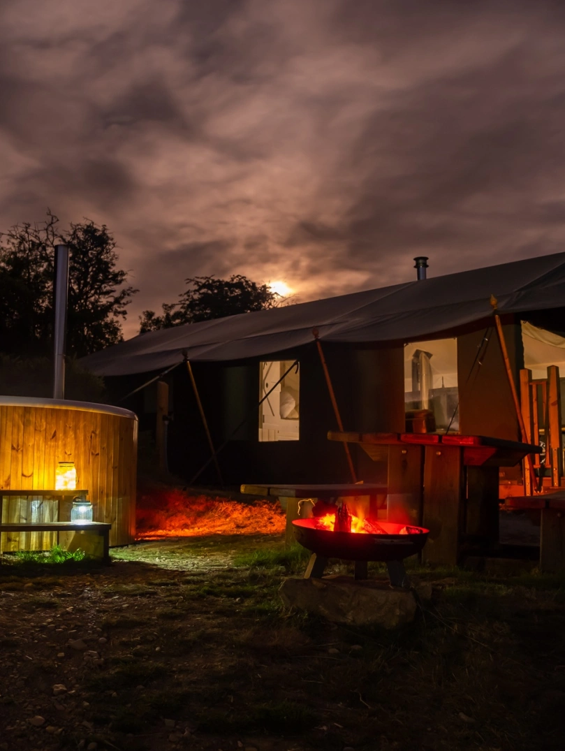 Night image of a glamping cabin, with a hot tub and lit firepit outside.