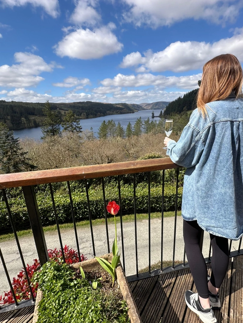 A lady holding a glass of fizz, on a balcony overlooking a lake.