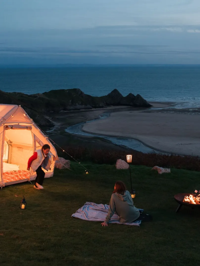 Family camping near a beach with a lighted tent and campfire.