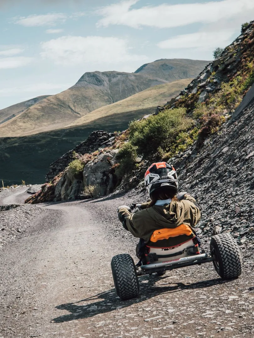 A person sitting in a go-kart travelling down a grey slate track with mountain views in the background.