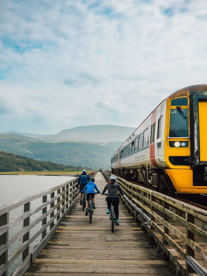 Three cyclists on a wooden railway bridge path, with a train passing by.