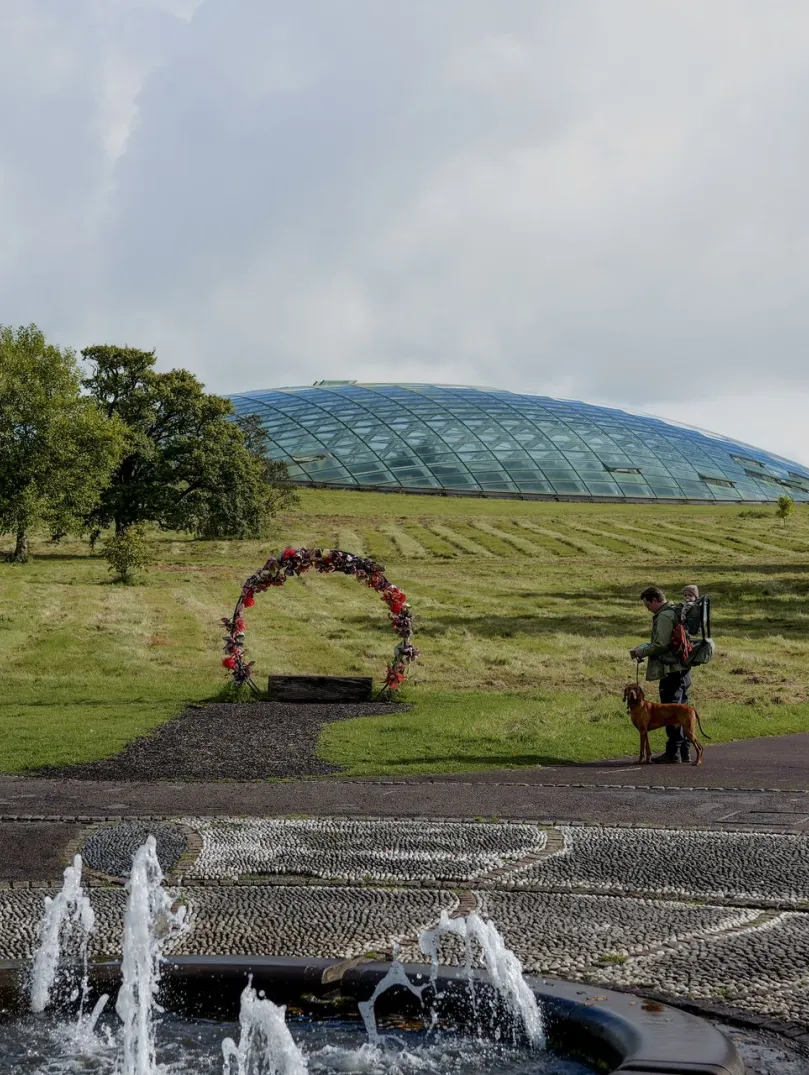 pond with water fountains, man with baby carried on his back and dog on lead with  great glasshouse in background.