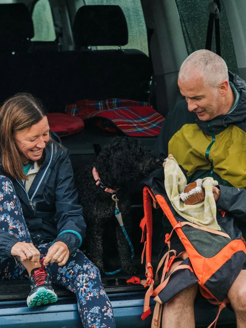 Couple sat in the back of a campervan putting on walking boots in preparation for a hike with their dog.