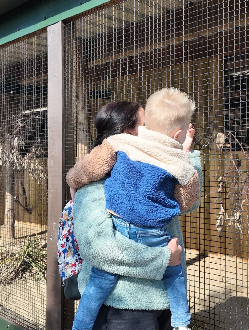 woman holding a child looks at a caged enclosure in a zoo, with some plants and branches inside.