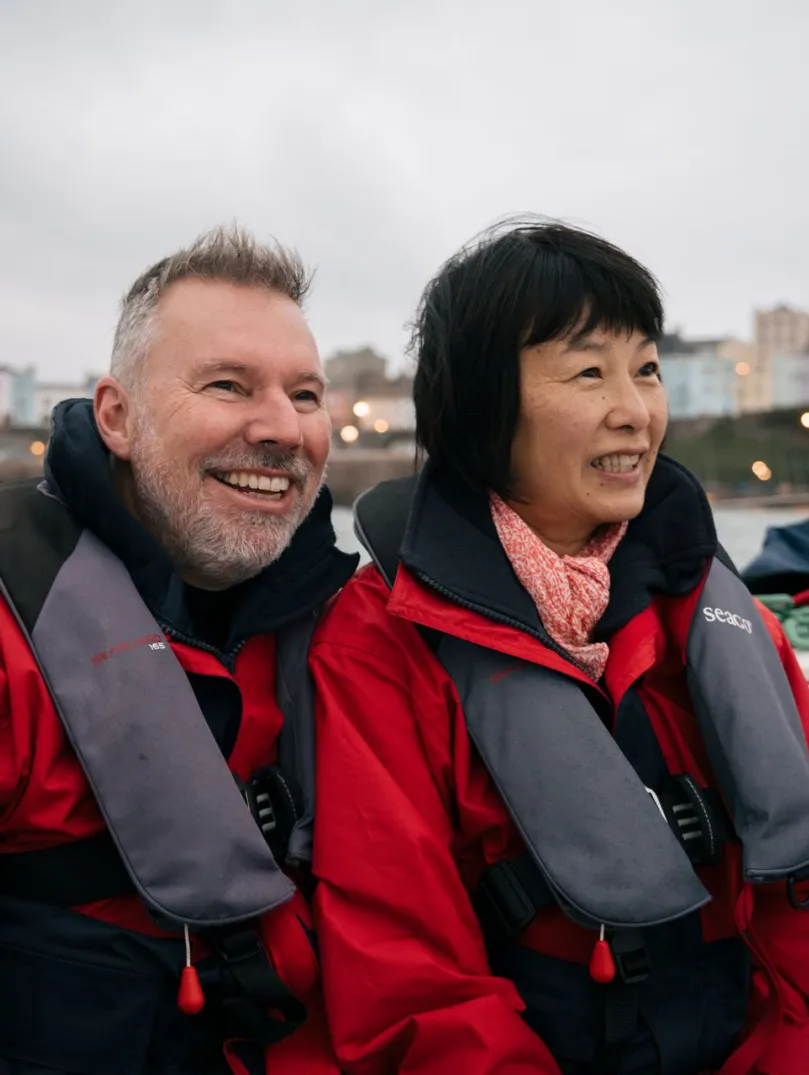 A man and woman wearing red life vests, on board a boat with Tenby harbour. in the background.
