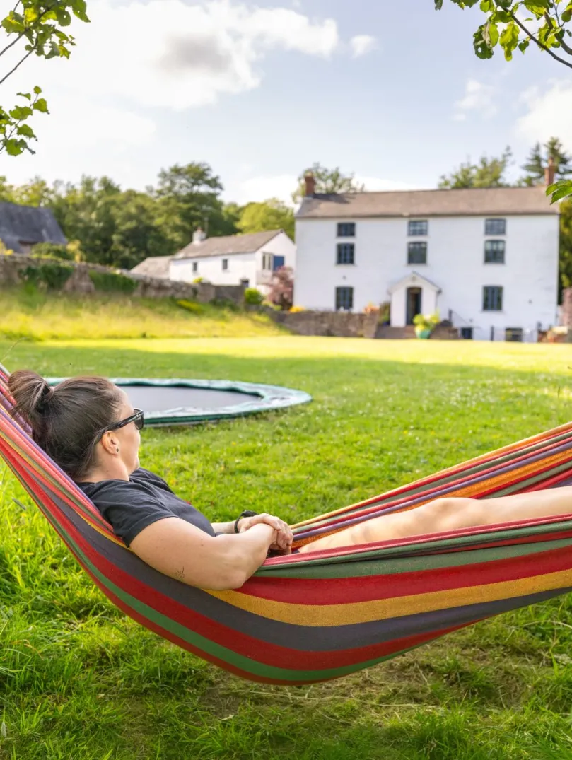 A person lying in a colourful hammock strung between two trees in a grassy garden, with a white farmhouse and trampoline in the background under a blue, partly cloudy sky