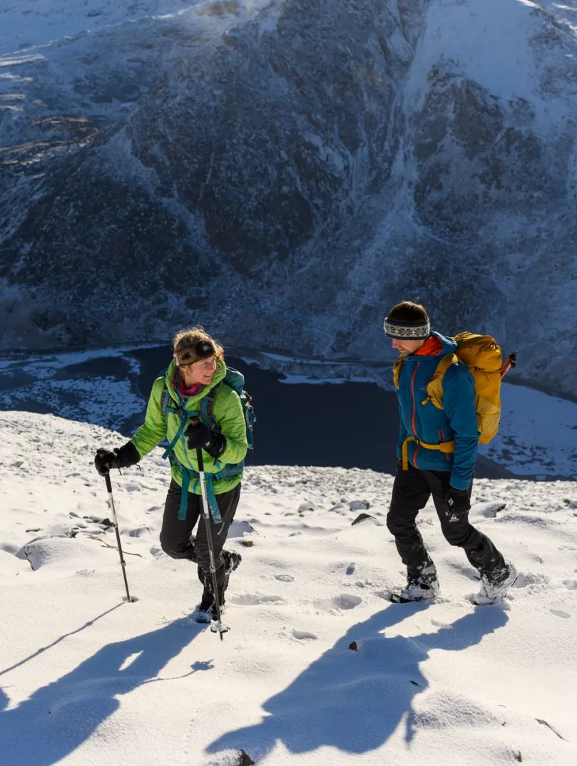 Two hikers walking on a snow-covered mountain summit with backpacks and poles, surrounded by rugged peaks under a partly cloudy sky.