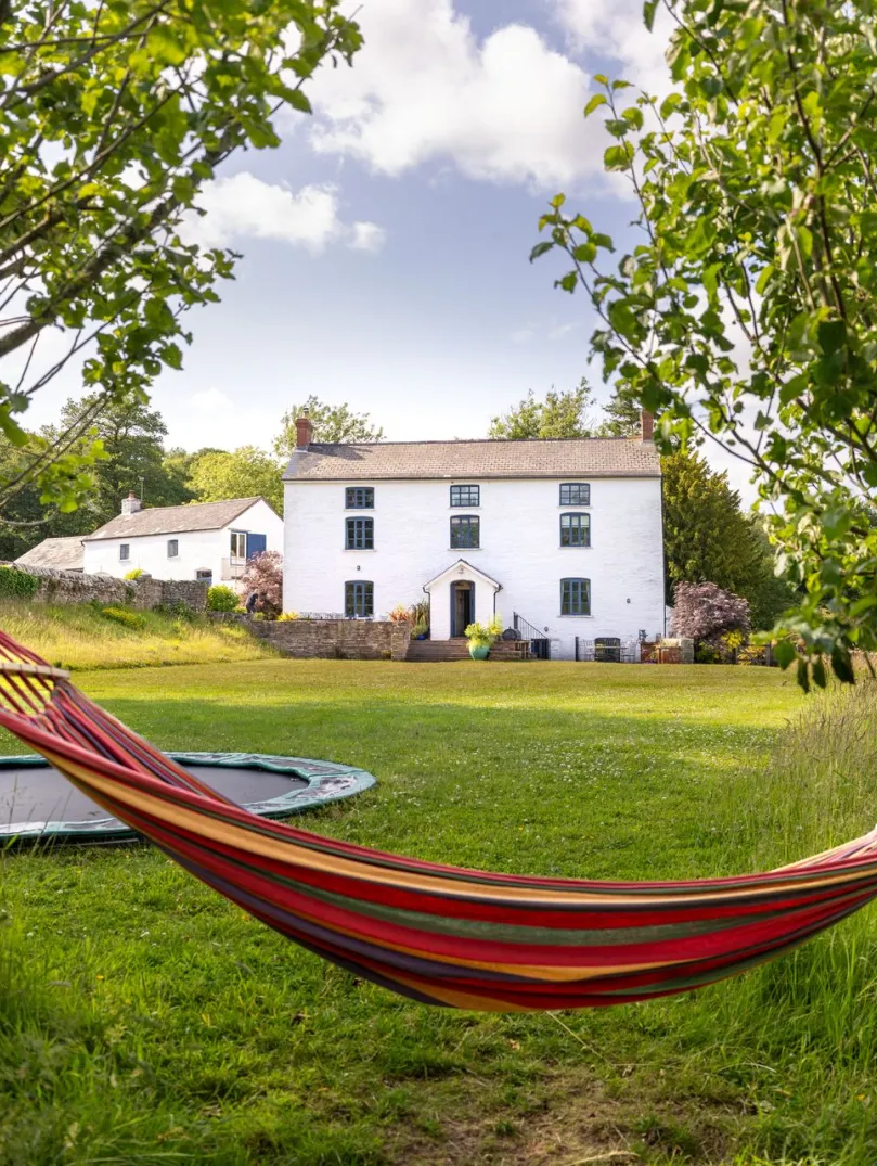 Colourful hammock strung between two trees in a grassy garden, with a white farmhouse and trampoline in the background under a blue, partly cloudy sky