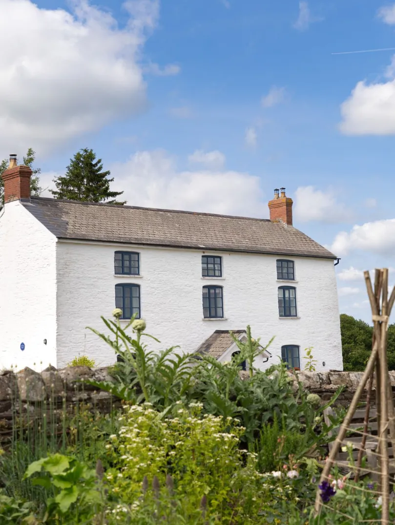 A large, white-washed cottage next to a flower garden. 