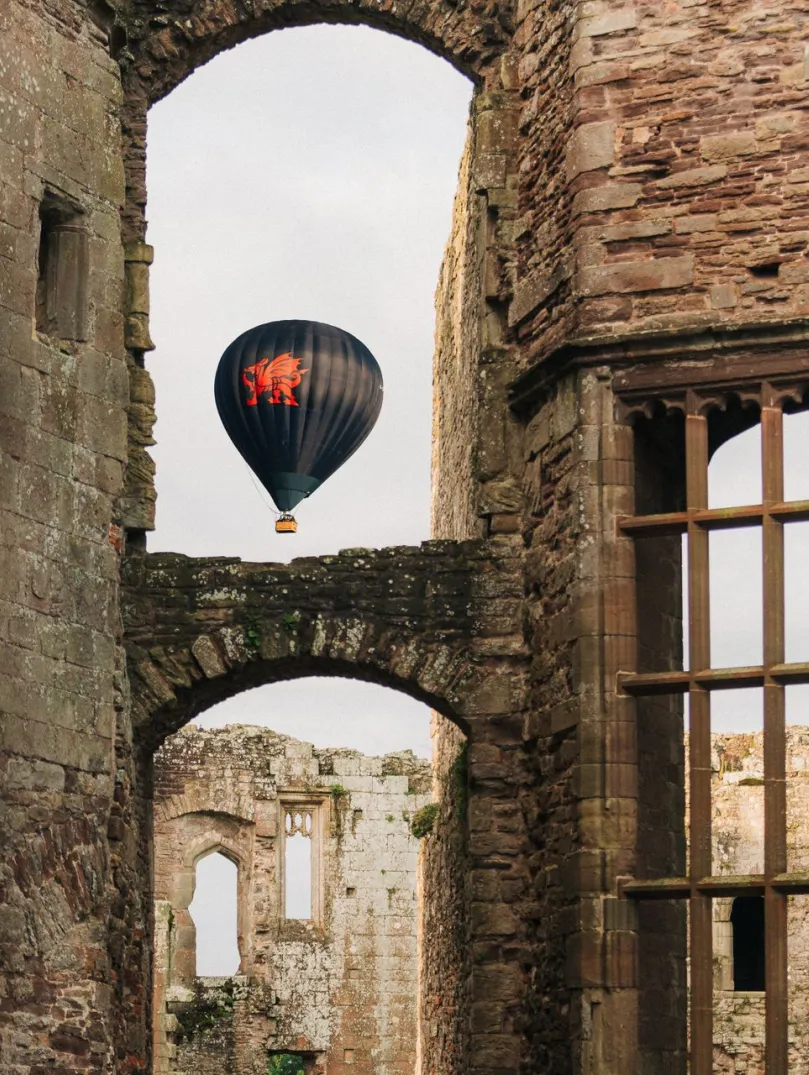 Hot air balloon with a red dragon design floating in the sky, framed by the stone arches and windows of a historic castle ruin.