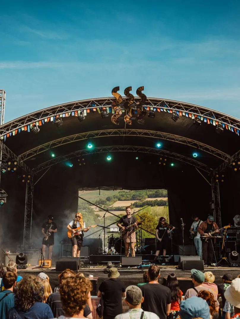 band playing at festival on a sunny day with crowd watching and hills in background.