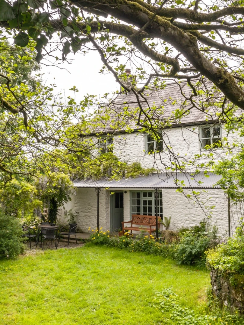 A white painted stone cottage, with a seating area outside, in a lawned garden.