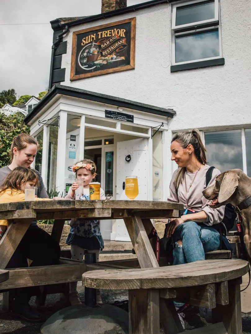 Family sit at a wooden picnic table outside a white pub with drinks, while two dogs stand nearby.