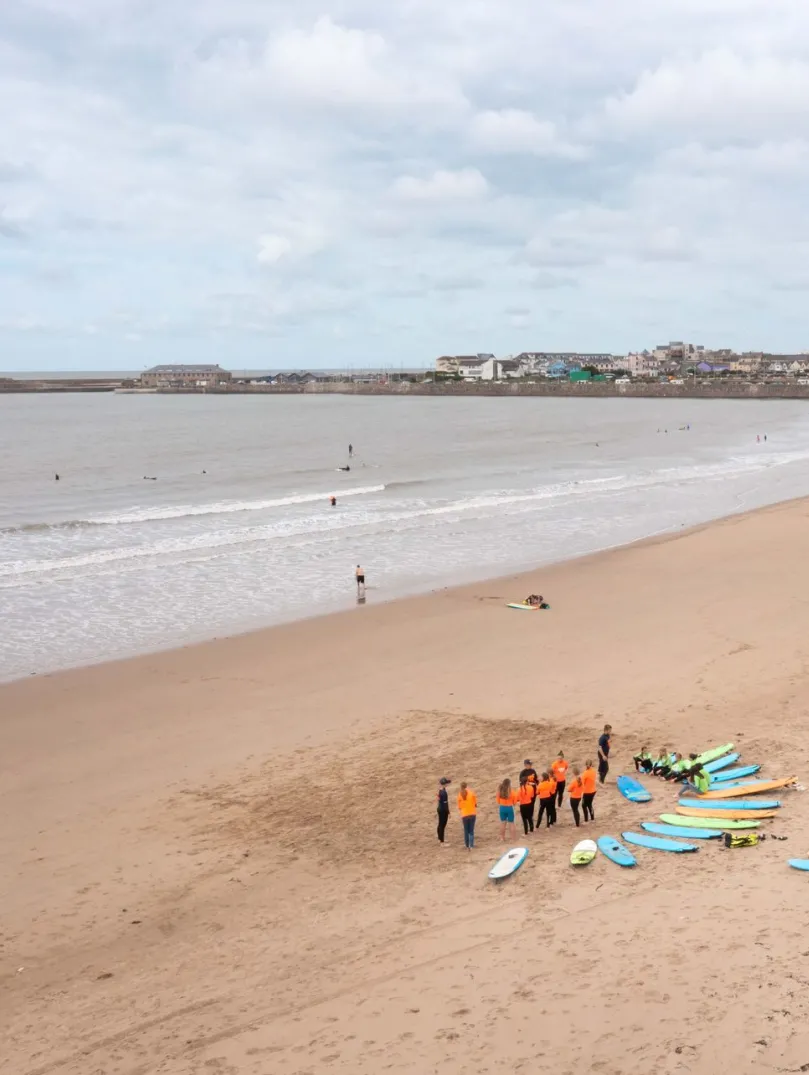 Surf lesson group with surfboards gathered on a wide sandy beach, with gentle waves and a coastal town in the distance.