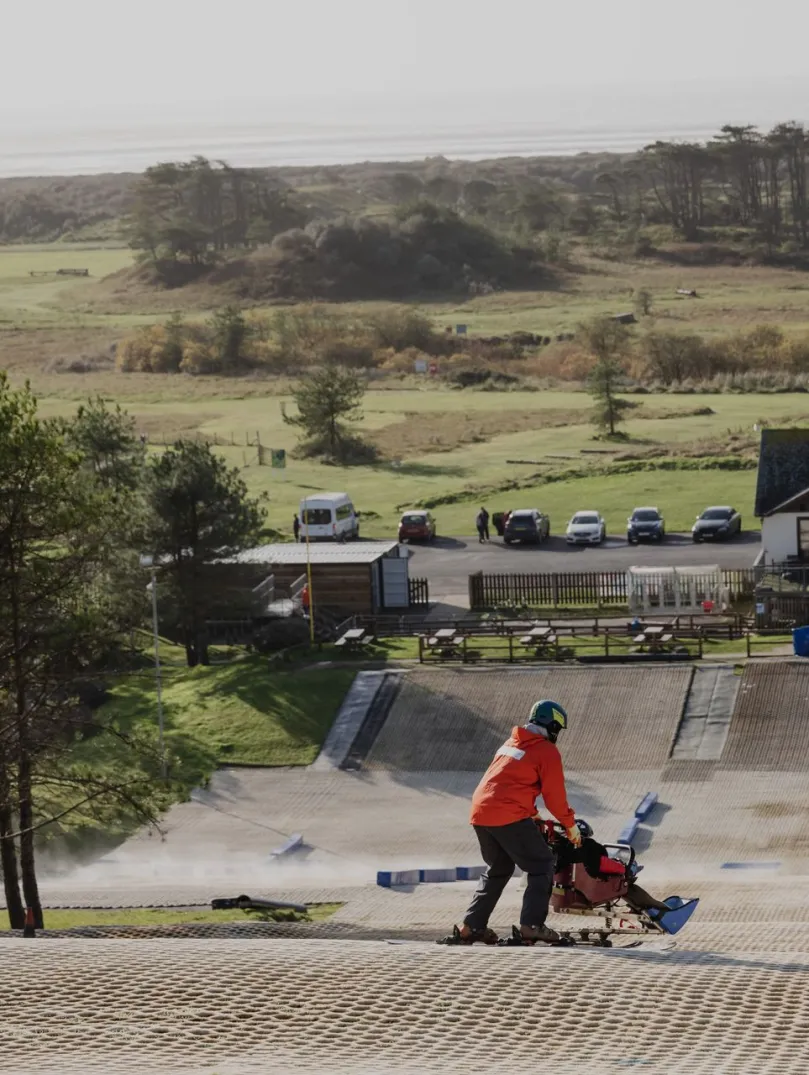 A child in an adapted sled / ski-pram being pushed by a man on a dry ski-slope.
