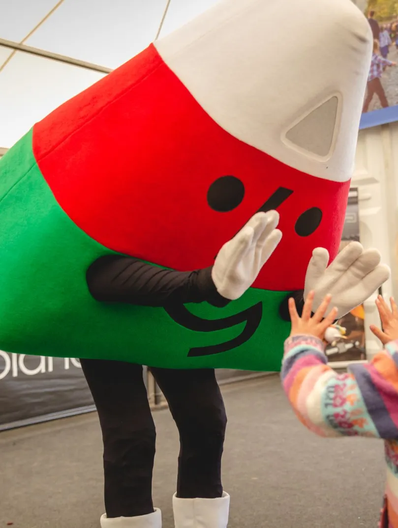 A costumed mascot in red and green leans down to greet a small child at an indoor event.