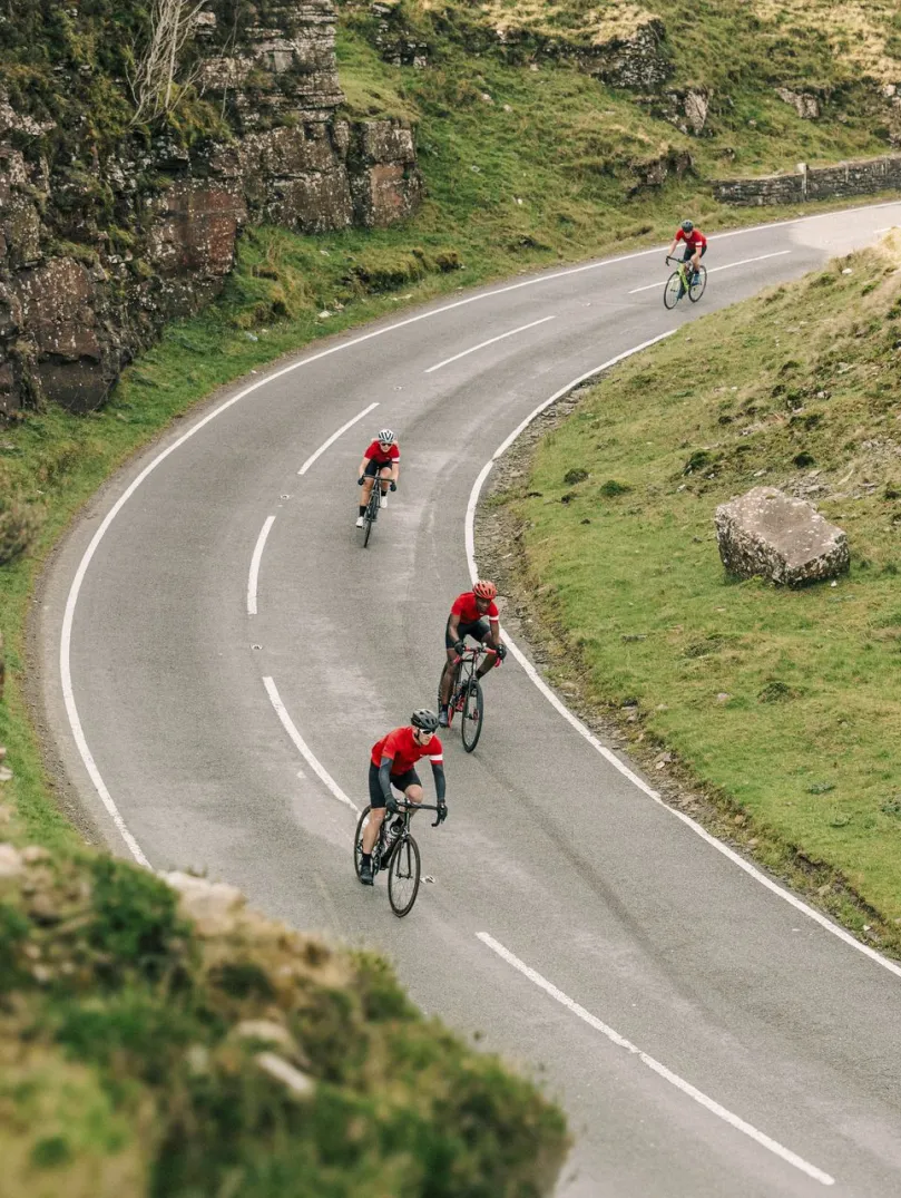 Cyclists climbing a winding mountain road past a rock face painted with a Welsh dragon and the words “Cymru am byth” and “...the Valleys”, surrounded by green hills.