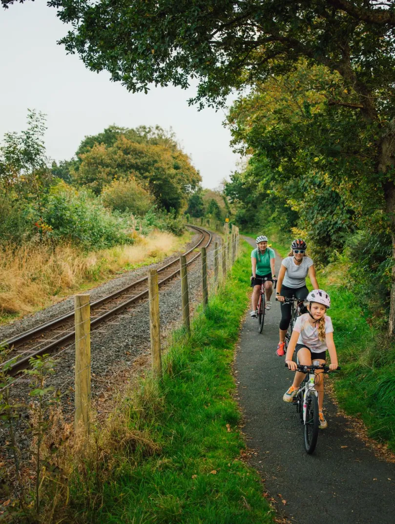Adult and child cycling along a narrow path beside railway tracks, with two more riders following under trees.