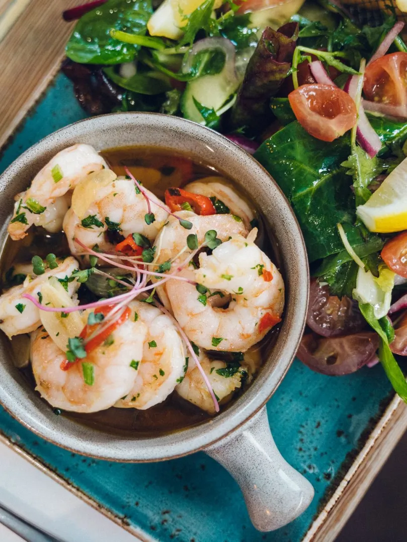 Bowl of prawns in a herb dressing served with mixed leaves, cherry tomatoes and a lemon wedge on a blue plate.