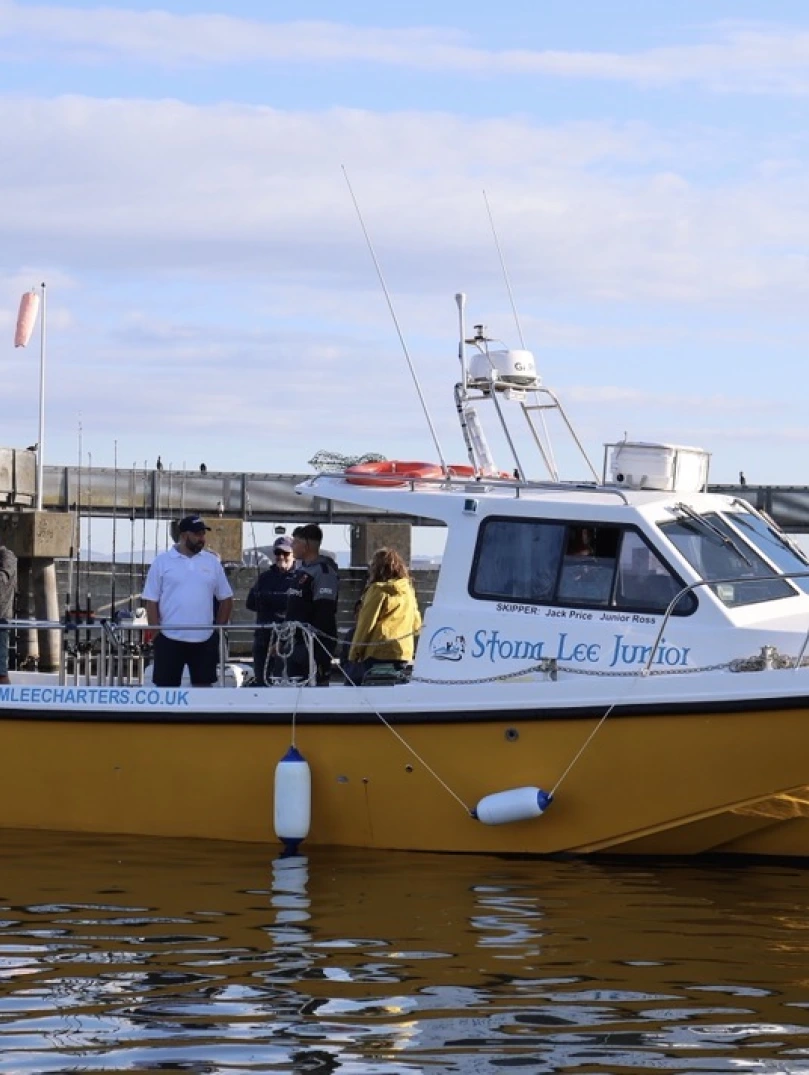 Passengers aboard the Storm Lee Junior charter boat on calm waters beside a harbour pontoon, with Cardiff Bay visible in the background under a partly cloudy sky.