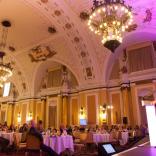 Delegates seated at round dining tables listening to a speaker in an ornate room.
