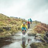 Three mountain bikers cycling downhill through a puddle on a purpose built dirt track.