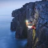 Two people sitting on a portaledge suspended off a cliff with the sea below