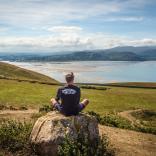 A man sitting on a rock overlooking some countryside and the coast.