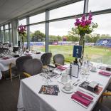 table decorated with white tablecoth and pink flowers for an event overlooking the cricket grounds with blue seats and trees in the background. 