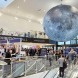 Exhibition hall with people coming up esculators to reception desks and a large grey space globe suspended from the ceiling.