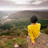 Woman in a yellow jumper sitting looking down into a partially forested, partially urbanised valley