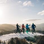 Four friends on mountain bikes on a snowy mountain top, looking out at the mountain scenery.