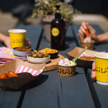 Food and drink on a black wooden bench. There are yellow coffee cups with the word BRAF on them, ice cream, strawberries, olives and nuts.