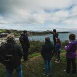 A tour guide with a group, pointing to a seaside town and castle from a cliff top.
