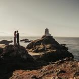 A wedding photograph of a couple kissing on top of a rock on a beach. There is a white lighthouse in the background.