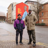 Two men in front of a mural on the side of a building. The mural shows a pregnant Black lady wearing a Cardiff City football shirt.