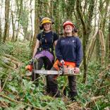 Two women in protective clothing holding chainsaws in a forest.