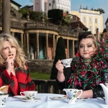 Two women having a cup of tea in ornate gardens.