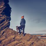 A person and a dog standing on rocks and looking out to sea.