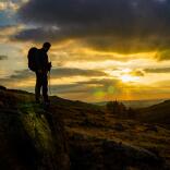Eine Person mit Rucksack, die in einer hügeligen Landschaft bei Sonnenaufgang steht.