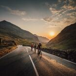 Three cyclists on a road with high mountains either side, at sunset.