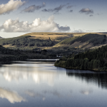Cloudy blue sky reflections in a flat reservoir, surrounded by woodland and mountains.