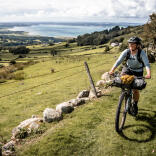 A female cyclist on a grassy roadway above a valley, looking down over the coast.