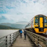 Three cyclists on a wooden railway bridge path, with a train passing by.