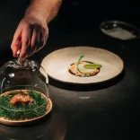 A person lifts a glass dome covering a dish with greens and a small item, next to a plate with arranged food on a dark surface.