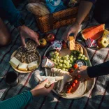 Bwyd picnic ar flanced. Ffrwythau, iogwrt a brechdan. Mae breichiau yn estyn allan am y wledd.