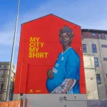 Mural painted on the gable end wall of a building, of a pregnant black woman wearing a colourful headscarf and blue football shirt cradling her baby bump.