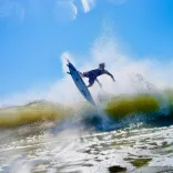 Surfer mid-air above a breaking wave, surrounded by water spray, under a bright blue sky.