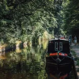 Black and red narrowboat moored on a tranquil canal lined with green trees, with people walking along the adjacent paved path.